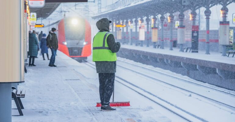 Ein Bahnsteig im Winter, bedeckt mit Schnee. Im Vordergrund steht eine Person mit einer neonfarbenen Warnweste und einem Besen, die den Schnee vom Bahnsteig fegt. Im Hintergrund nähert sich ein roter Zug dem Bahnhof, während andere Fahrgäste auf den Zug warten. Es schneit stark, und die Sicht ist durch den Schneefall eingeschränkt.