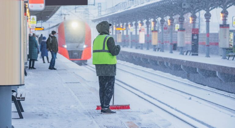 Ein Bahnsteig im Winter, bedeckt mit Schnee. Im Vordergrund steht eine Person mit einer neonfarbenen Warnweste und einem Besen, die den Schnee vom Bahnsteig fegt. Im Hintergrund nähert sich ein roter Zug dem Bahnhof, während andere Fahrgäste auf den Zug warten. Es schneit stark, und die Sicht ist durch den Schneefall eingeschränkt.